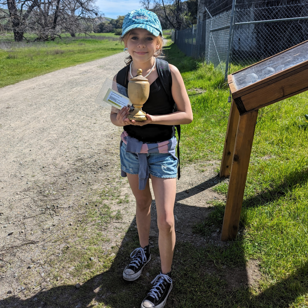 A smiling child outdoors holding a trophy and a certificate, standing beside a trail with greenery.