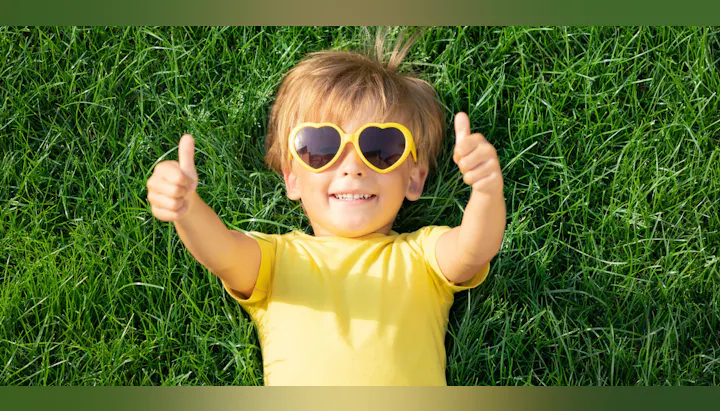 A cheerful child in a yellow shirt and heart-shaped sunglasses lies on lush green grass, giving two thumbs up.