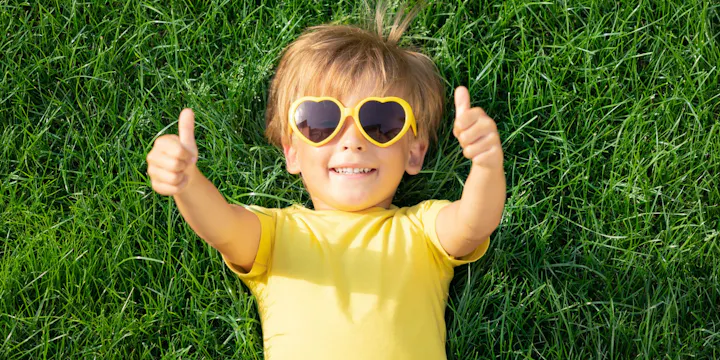 A cheerful child in a yellow shirt and heart-shaped sunglasses lies on lush green grass, giving two thumbs up.