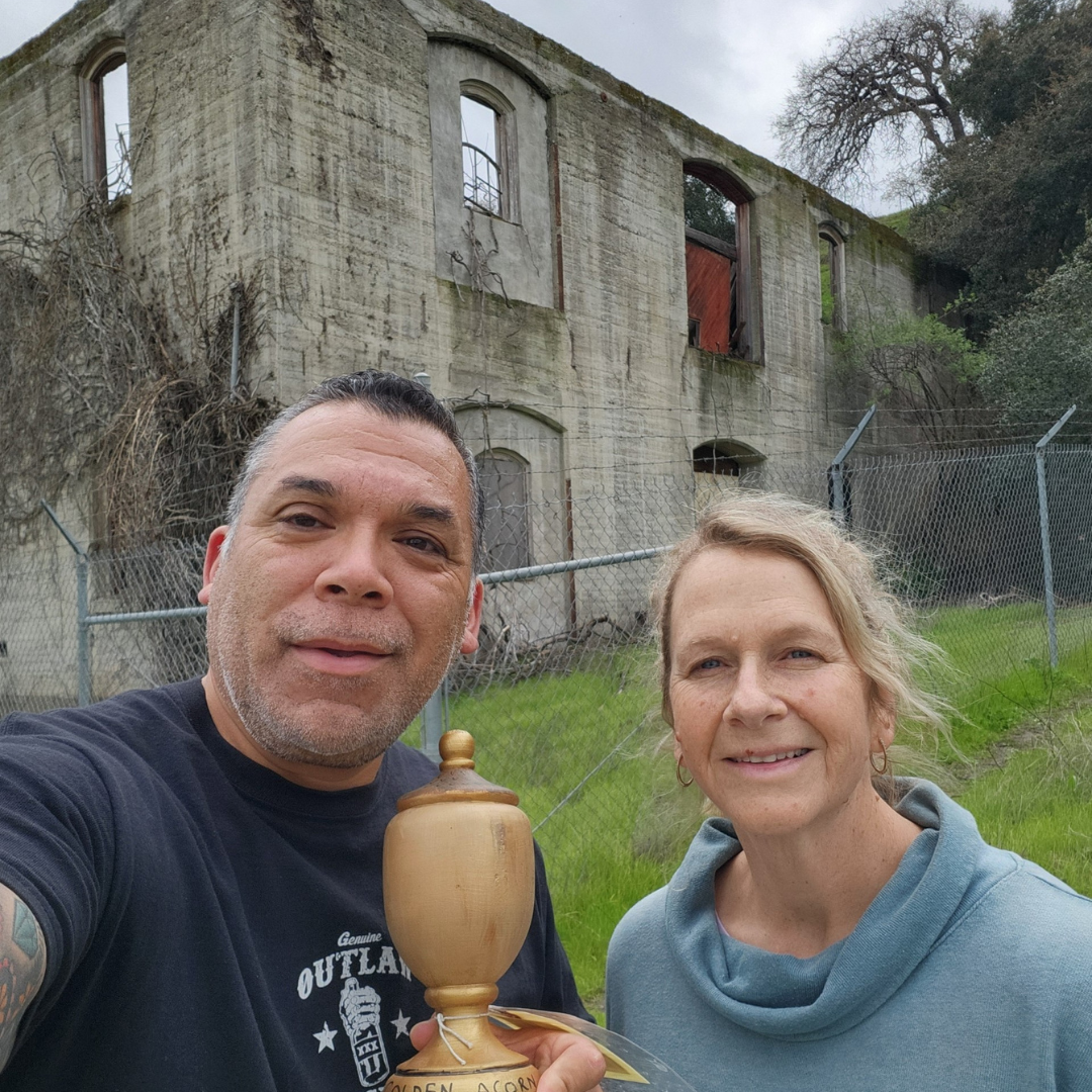 Two people holding a wooden object with a dilapidated building in the background.