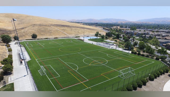 A multi-purpose sports field with goals and colored lines, surrounded by trees and hills, under a clear sky.