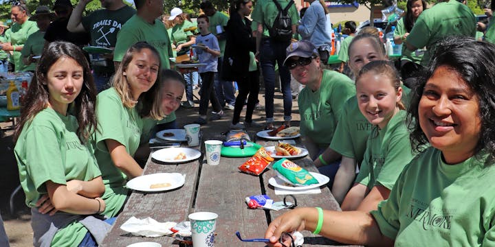 A group of people in green shirts sitting at a picnic table with food and drinks, surrounded by a crowd outdoors.