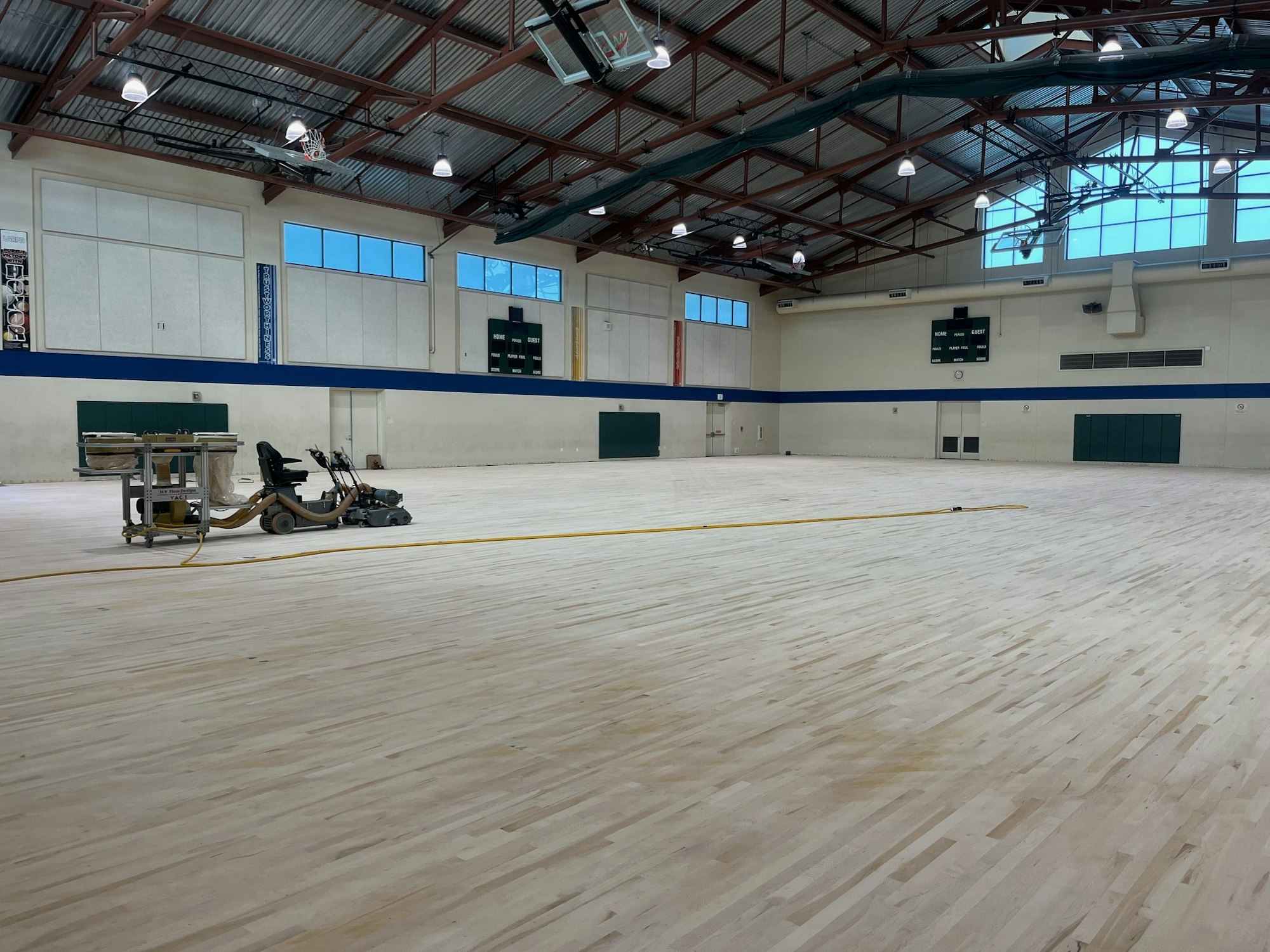 A gymnasium with light-colored wooden floors being sanded. A sanding machine is in use. High ceilings and scoreboard visible.