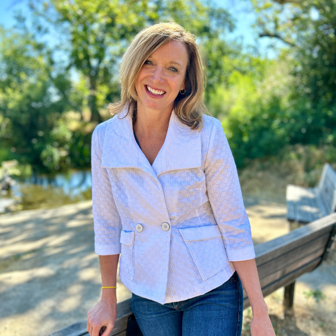 A person in a white jacket smiling, standing outdoors near a bench and trees in the background.