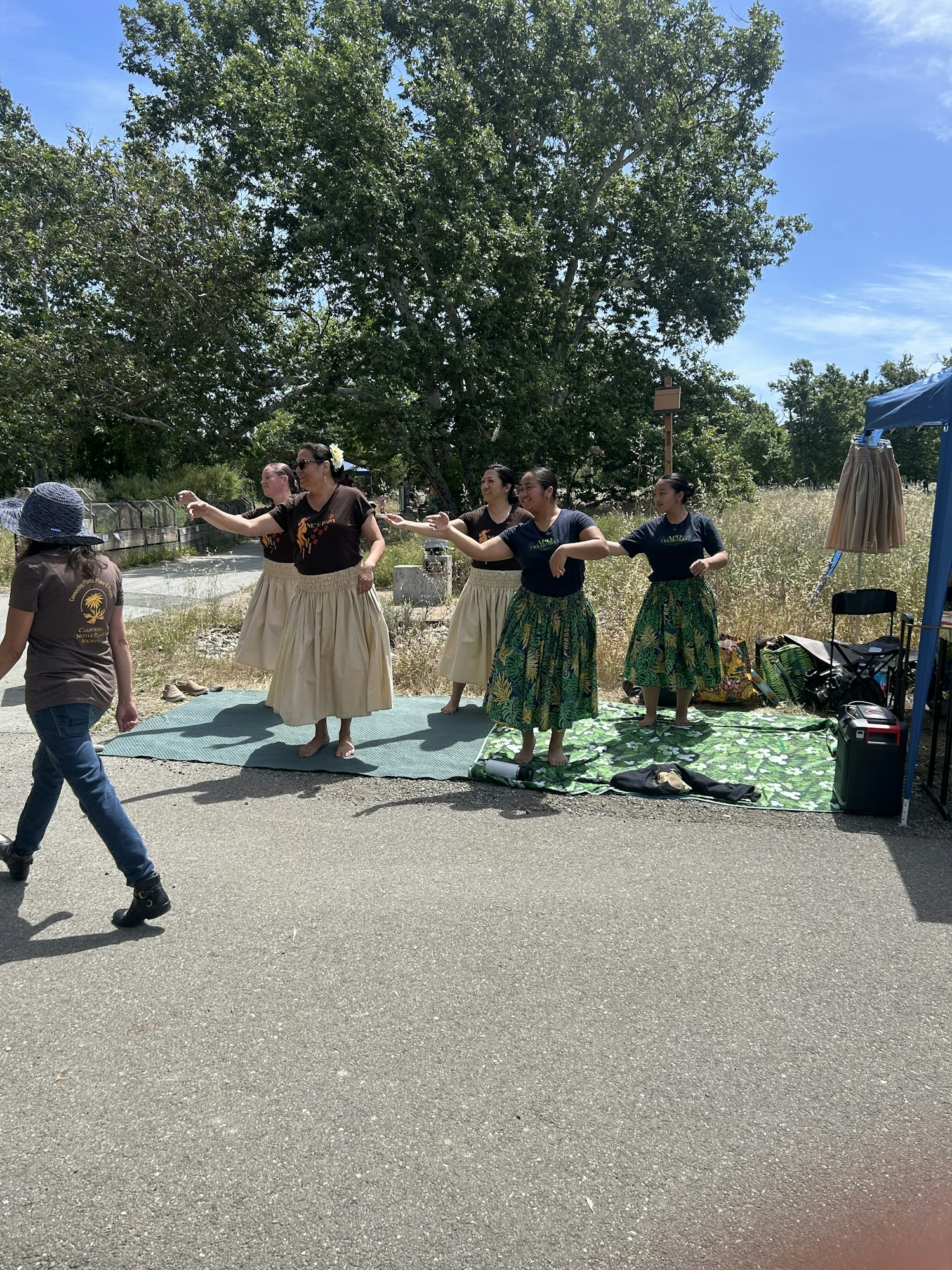 A group of dancers in traditional attire performing outdoors, with a tree and a clear blue sky in the background.