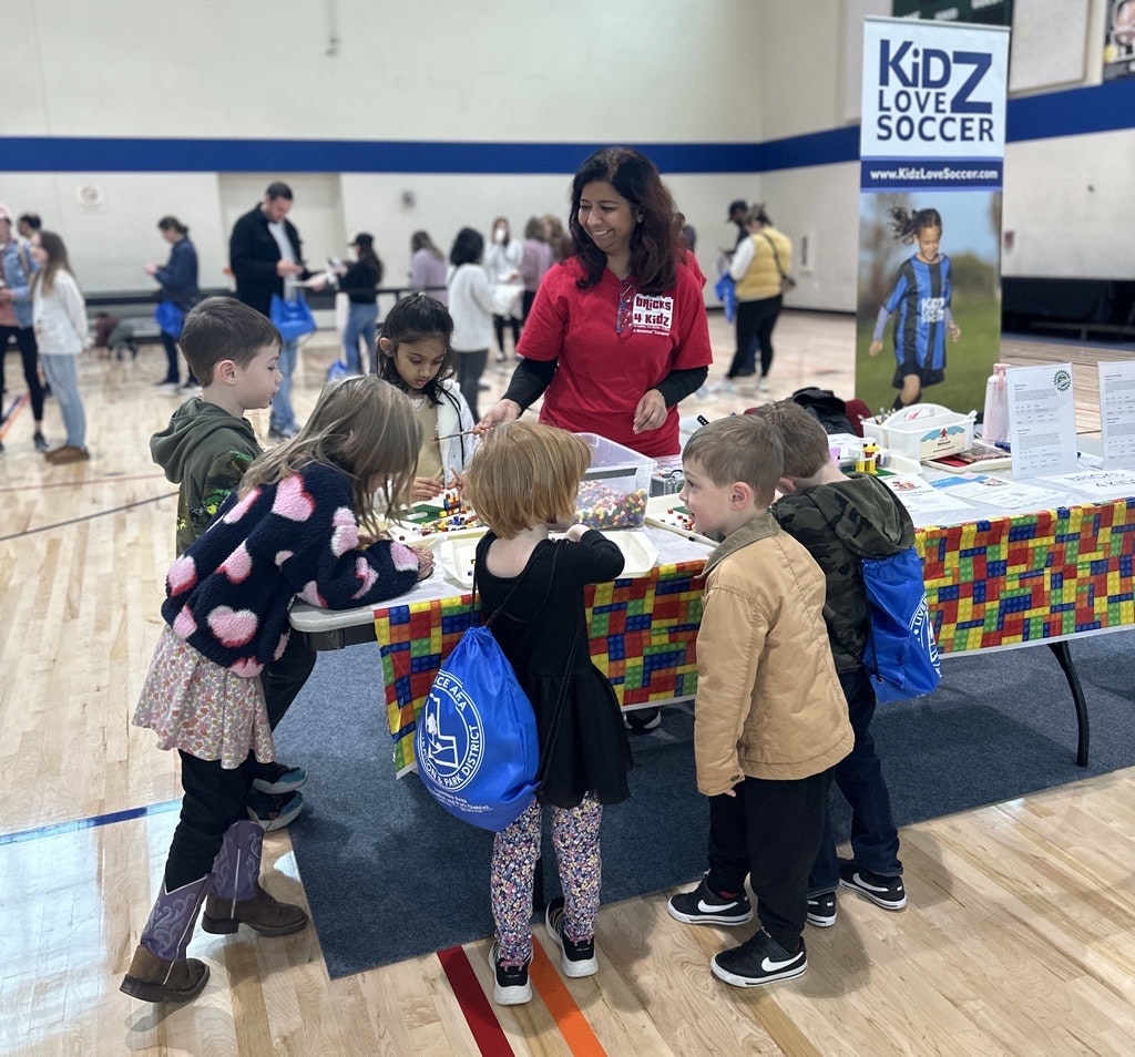A group of children is gathered around a table with activities while an adult assists them, promoting a soccer program.