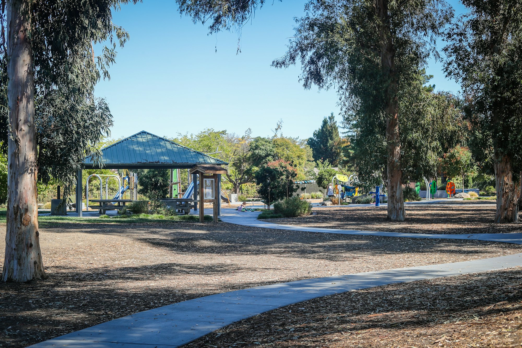 A sunny park with trees, a gazebo, and playground equipment in the background, with walking paths and people enjoying the area.