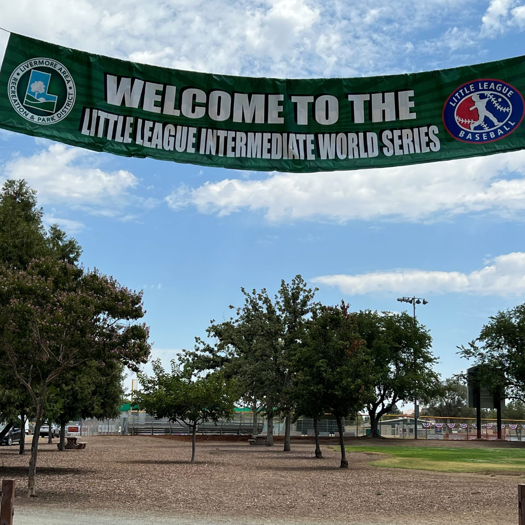 A banner saying "WELCOME TO THE LITTLE LEAGUE INTERMEDIATE WORLD SERIES" in a park with trees and benches.