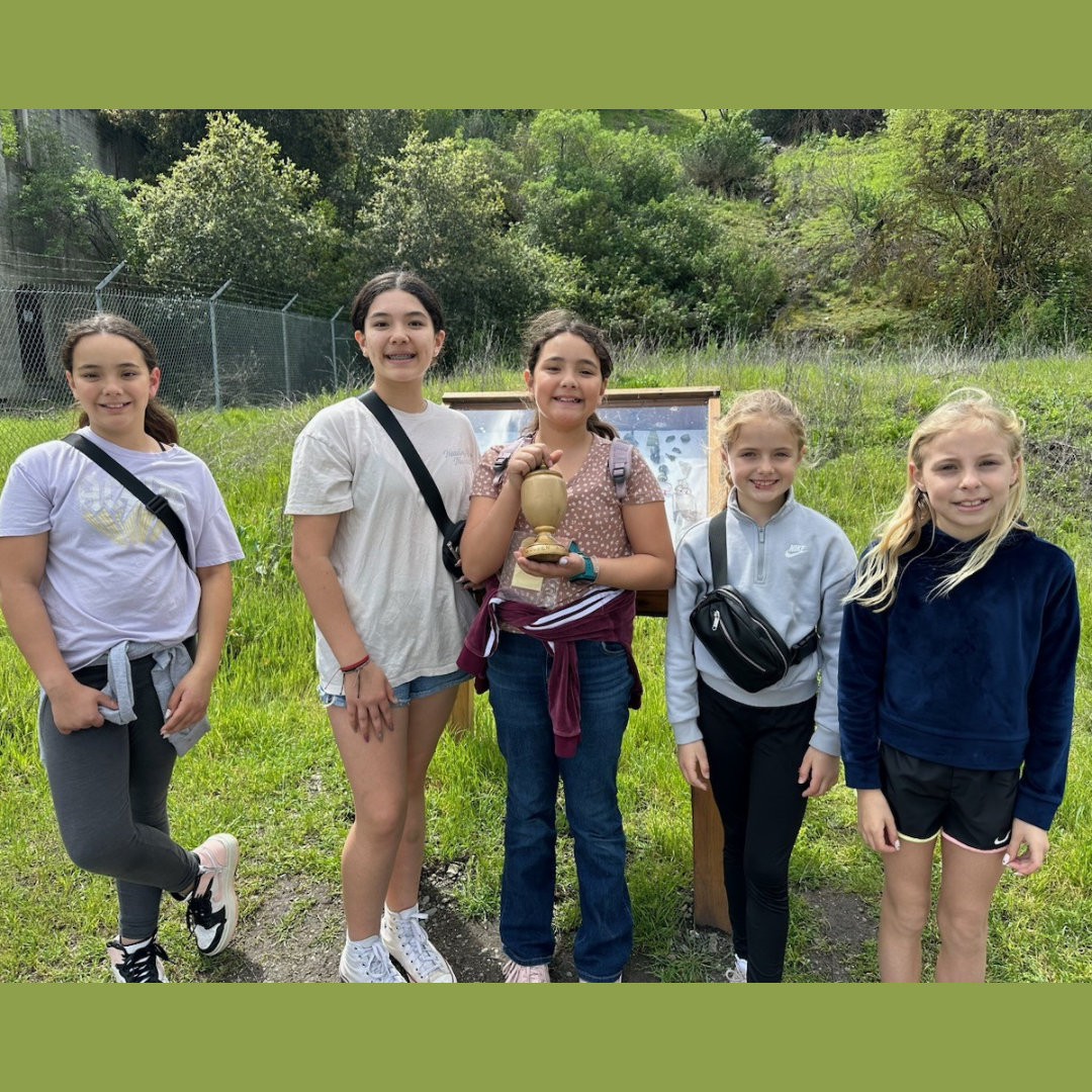 Five smiling kids outdoors, one holding an object, with greenery in the background.