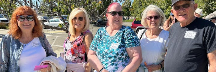 A group of five people smiling outdoors, wearing sunglasses and name tags, with cars and trees in the background.