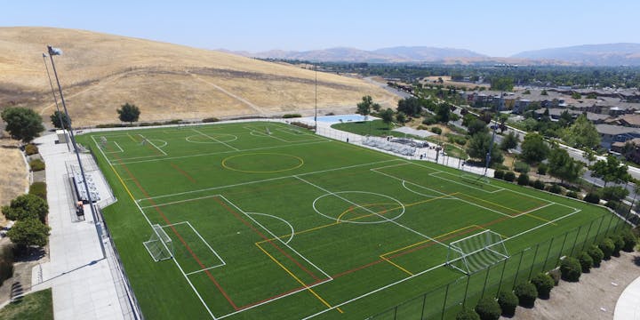 Aerial view of a large multi-sport field with various markings, surrounded by hills, pathways, and residential areas.