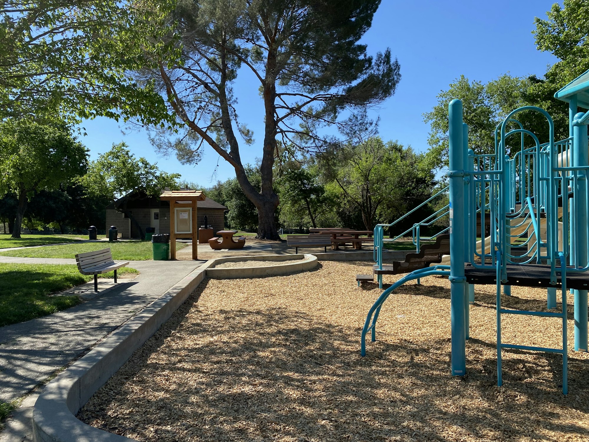 A playground with a slide and climbing structure, a bench, and lush trees in a park setting.