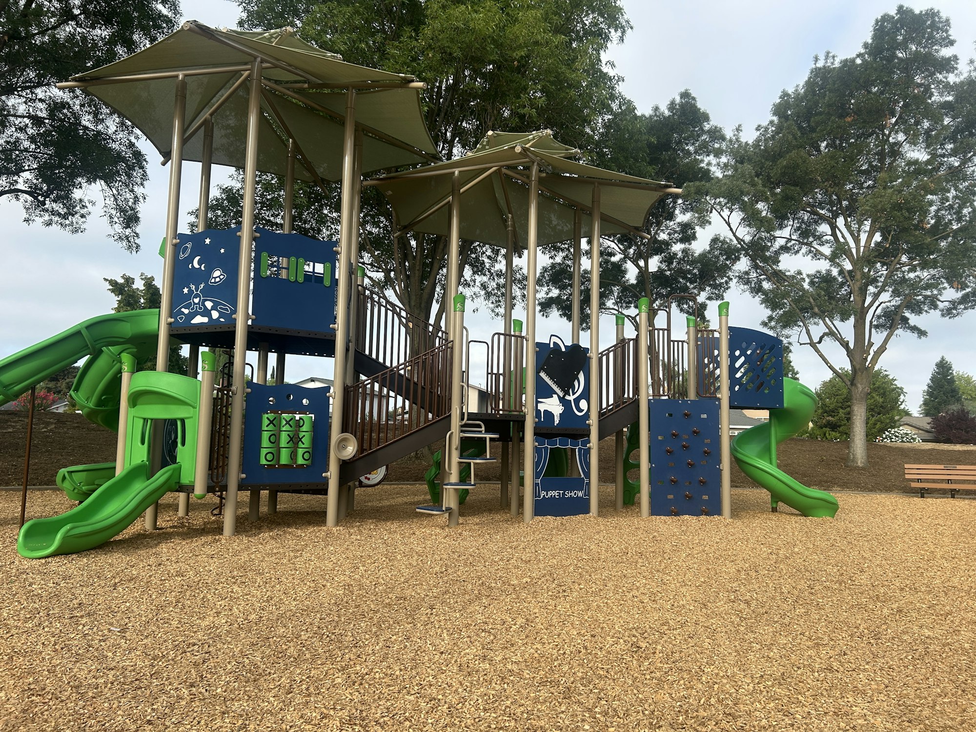 Playground with slides, climbing walls, tic-tac-toe, and a puppet show area, surrounded by trees and woodchip ground.