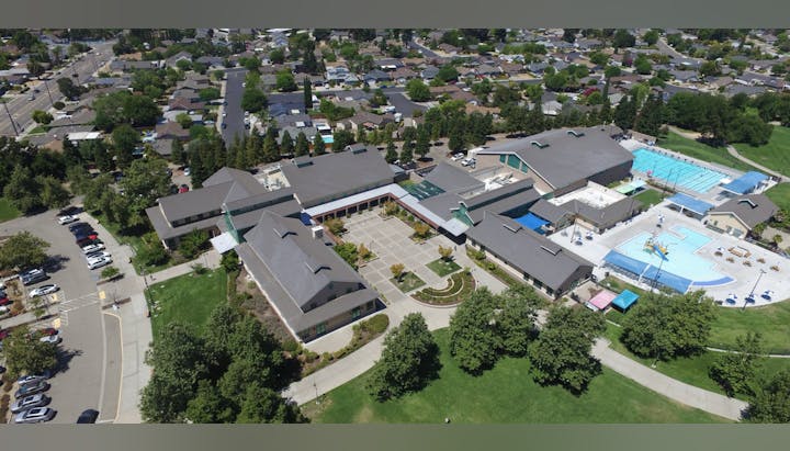 Aerial view of a large building complex with parking, swimming pools, and nearby residential area.