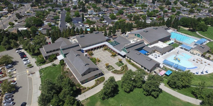 Aerial view of a large building complex with parking, swimming pools, and nearby residential area.
