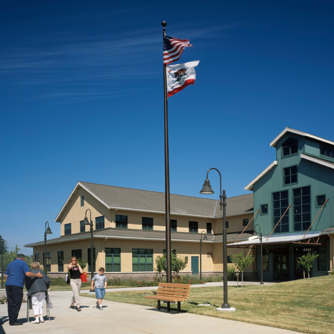 A modern building with people walking nearby, two flags flying on a pole, a pathway, and a bench in a landscaped area.