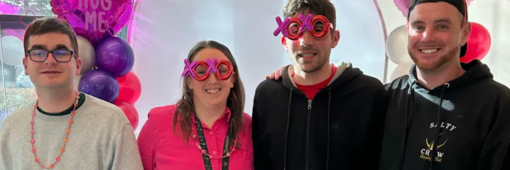 Four people smiling, two wearing "XOXO" glasses, in front of colorful balloons and a "HUG ME" balloon.