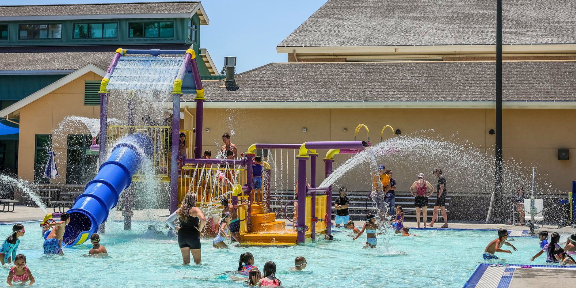 A lively water park scene with children playing, a large water slide, and splashing water features on a sunny day.