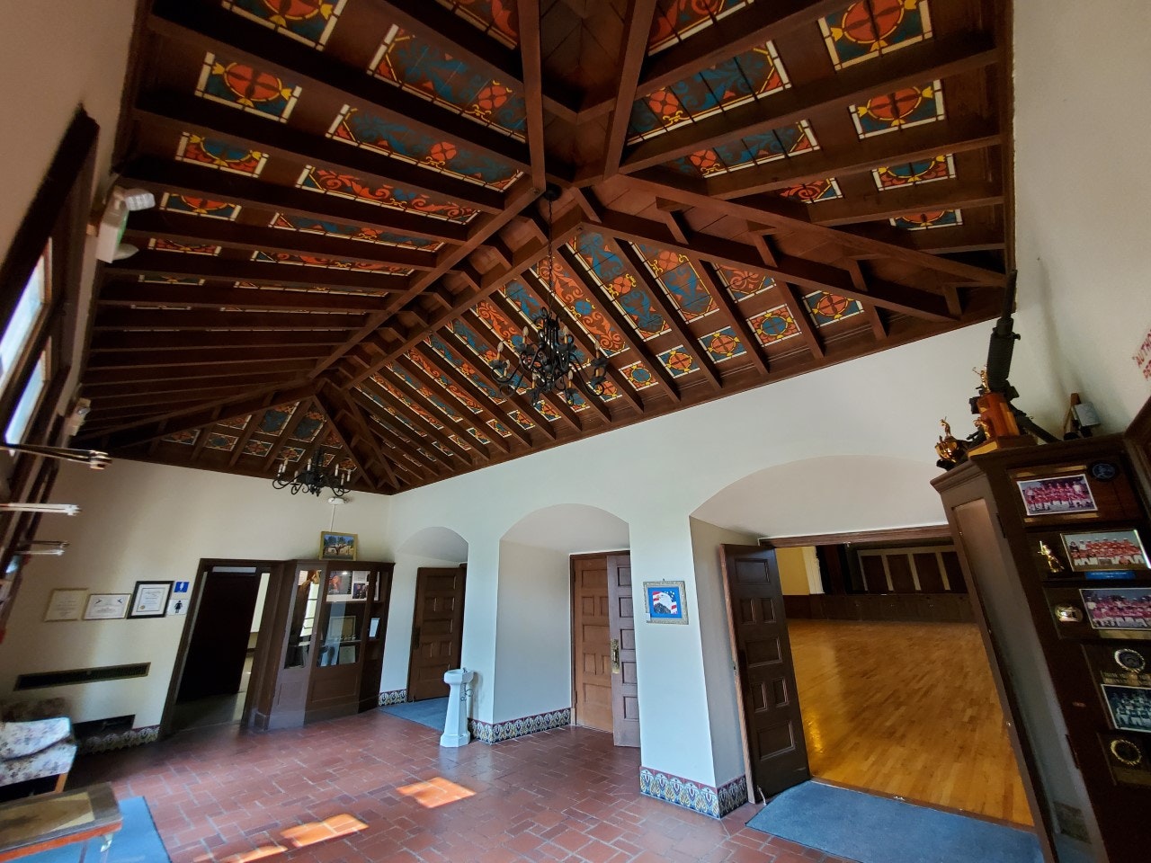 A room with a wooden ceiling, colorful stained-glass details, doors, plaques, and trophies on a cabinet.