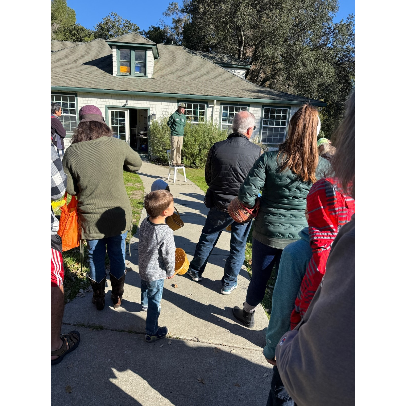 A group of people gather outside a house, listening to someone speaking from a stool.