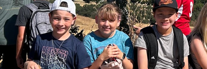 Three kids smiling outdoors, wearing casual clothes and backpacks, with trees and dry grass in the background.