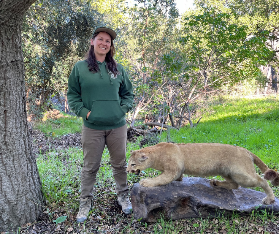 A person standing next to a young lion in a natural setting.