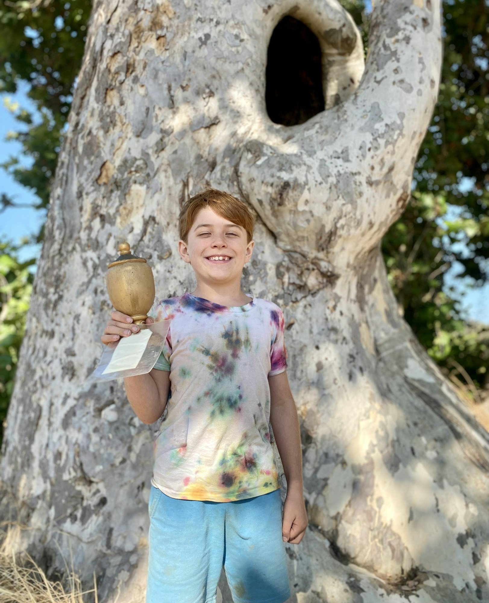 A smiling child holding a trophy stands in front of a tree with a large hollow.
