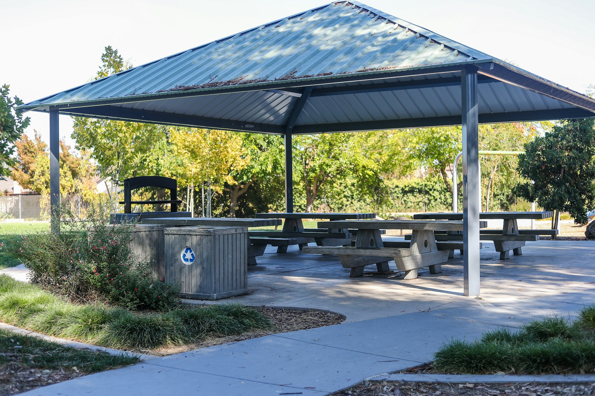 A covered picnic area with tables, a trash bin, and greenery in a park.