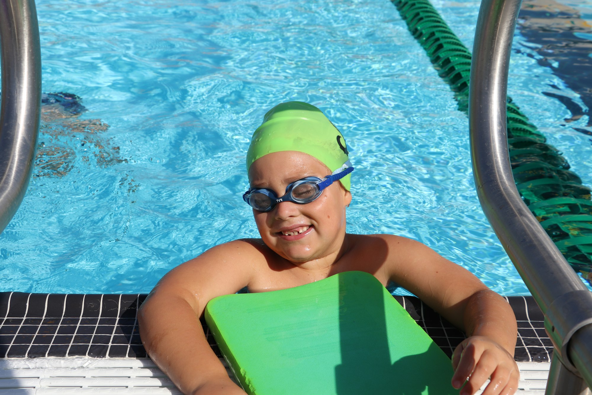 Child in a pool wearing a green swim cap and blue goggles, holding onto a green kickboard.