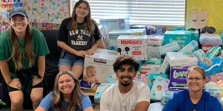 A group of smiling people poses with a large collection of diapers and baby supplies in a colorful room.