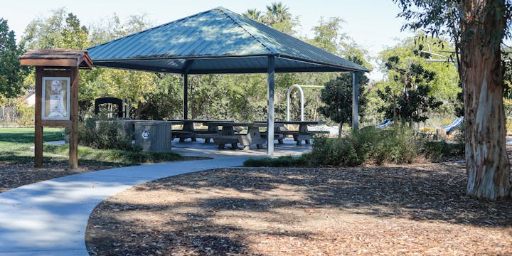 Park pavilion with benches, a notice board, and trees. Pathway leads to play area with slides.