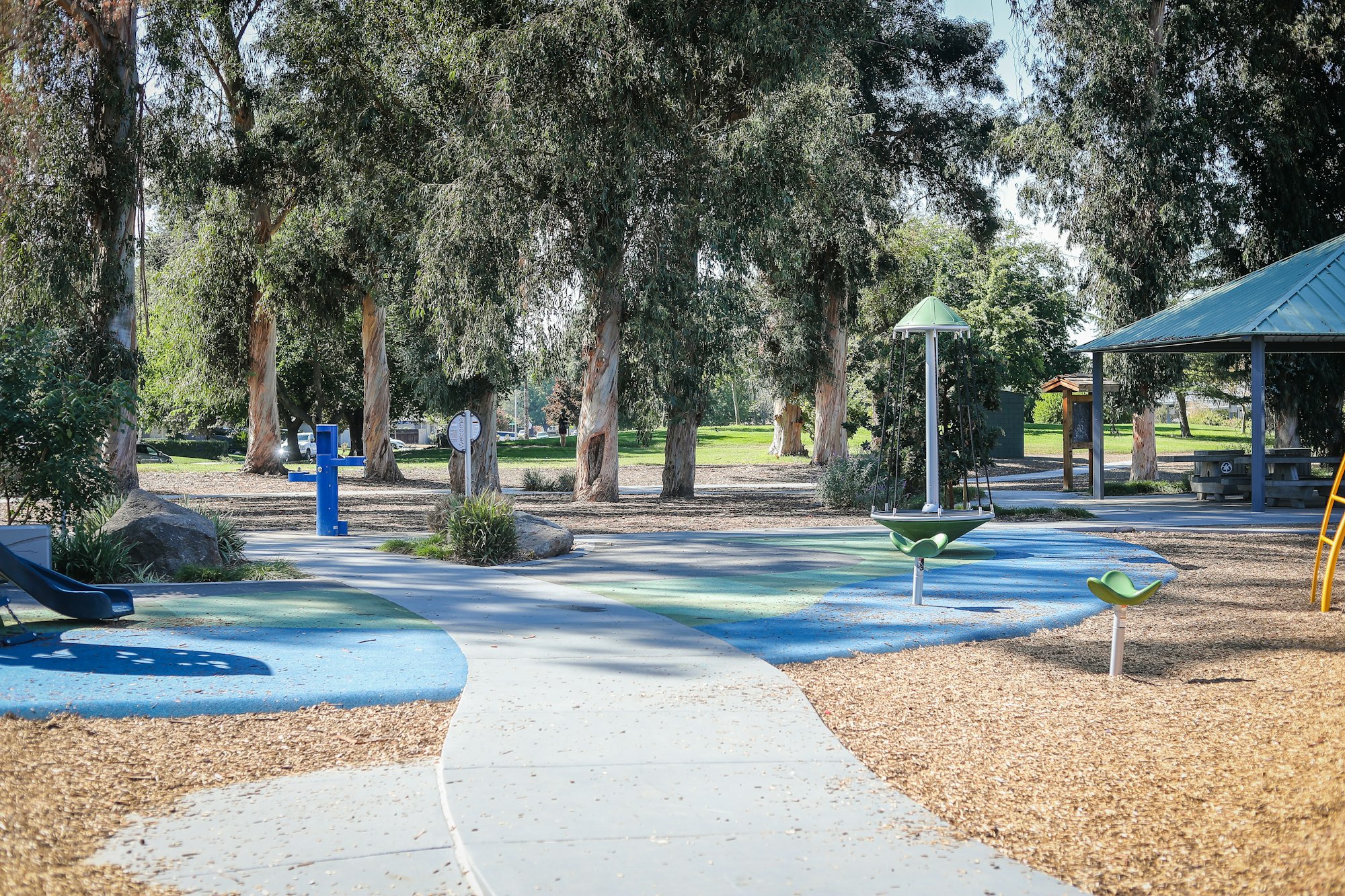 A playground with colorful ground, trees, spinning equipment, a shelter, and a walking path.