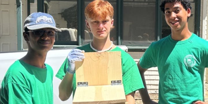 Three people in green shirts, one holding a wooden object with hinges, smiling outdoors.