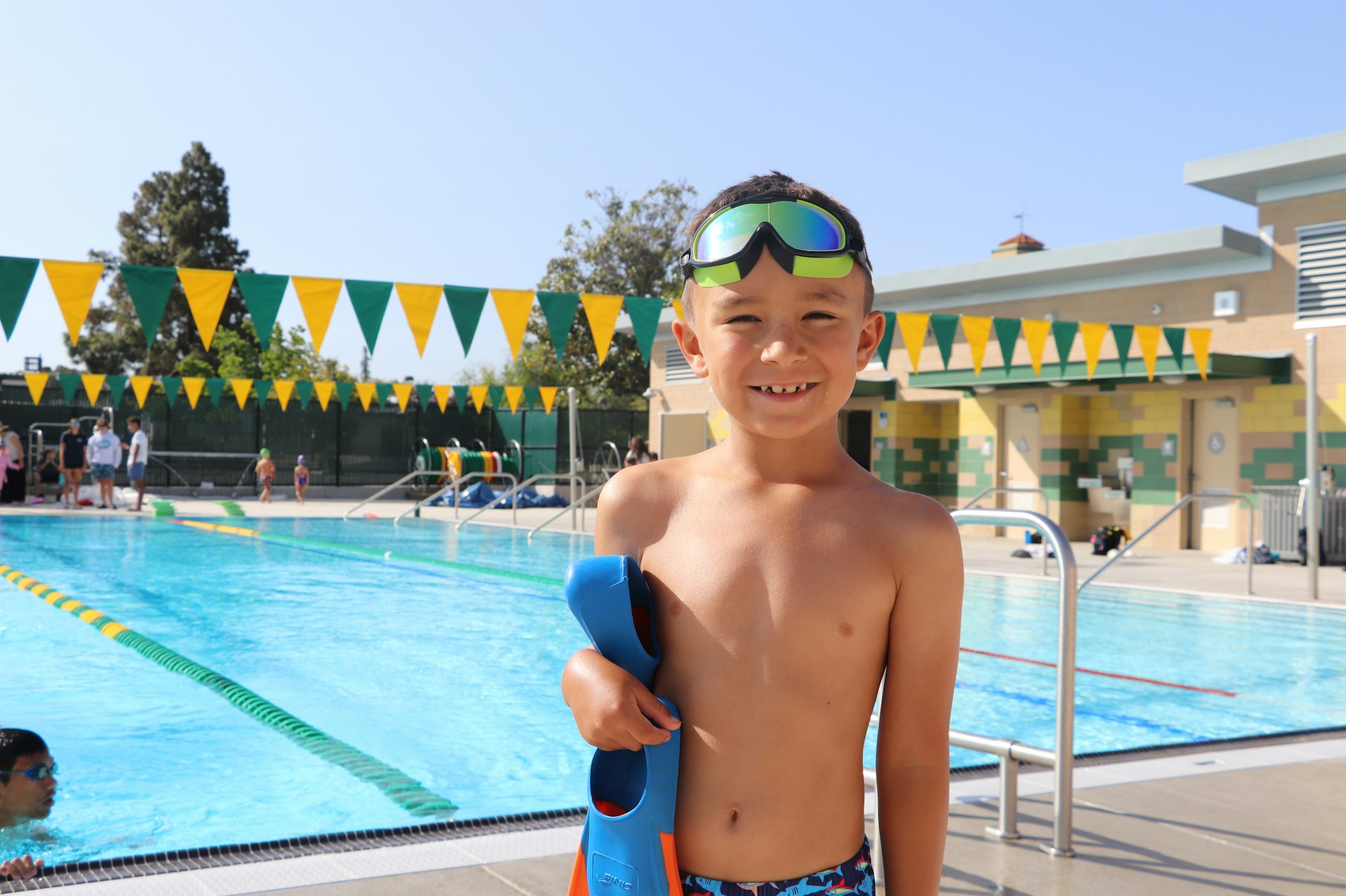 A child with goggles and flippers stands by a pool, smiling. Green and yellow flags adorn the background.