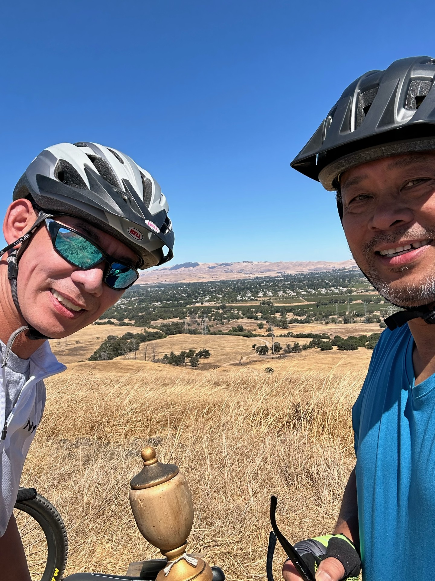 Two people in cycling helmets taking a selfie with a trophy and a scenic landscape in the background.