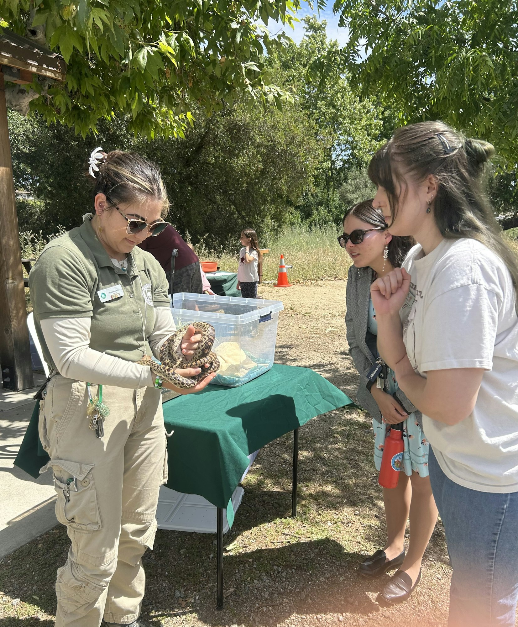 A nature event featuring a woman holding a snake, while two spectators watch and engage with curiosity outdoors.