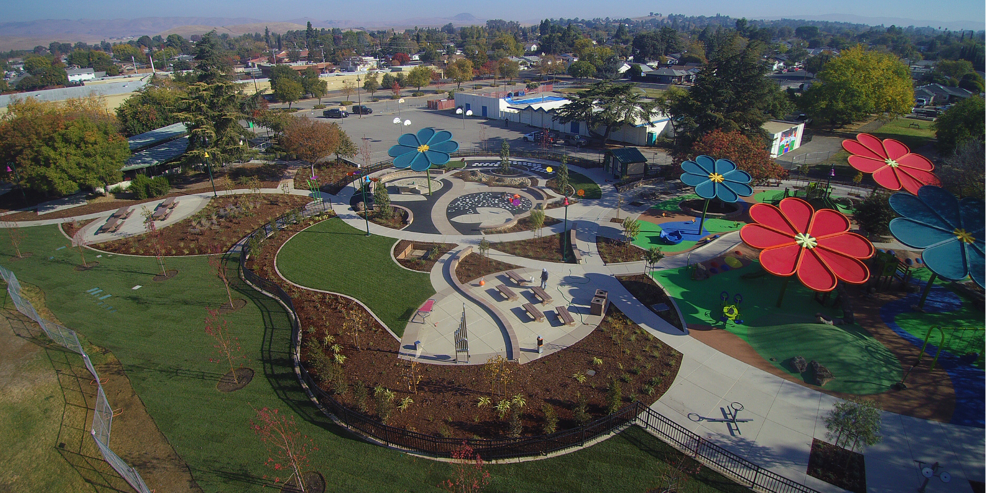 Colorful playground with large flower structures, winding pathways, and seating areas, surrounded by greenery and trees.