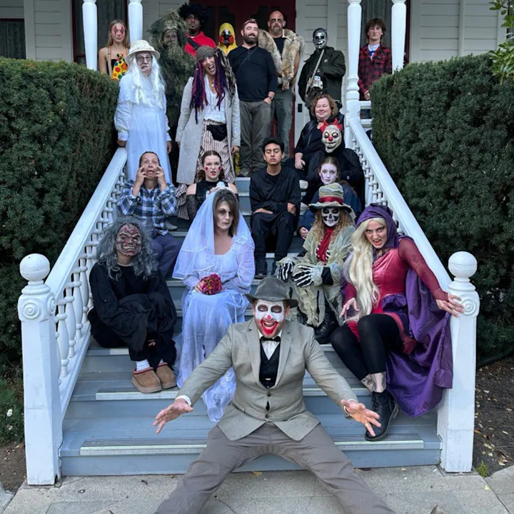 A large group in costumes, including ghosts, clowns, and zombies, poses on the stairs of a house for a Halloween-themed photo.