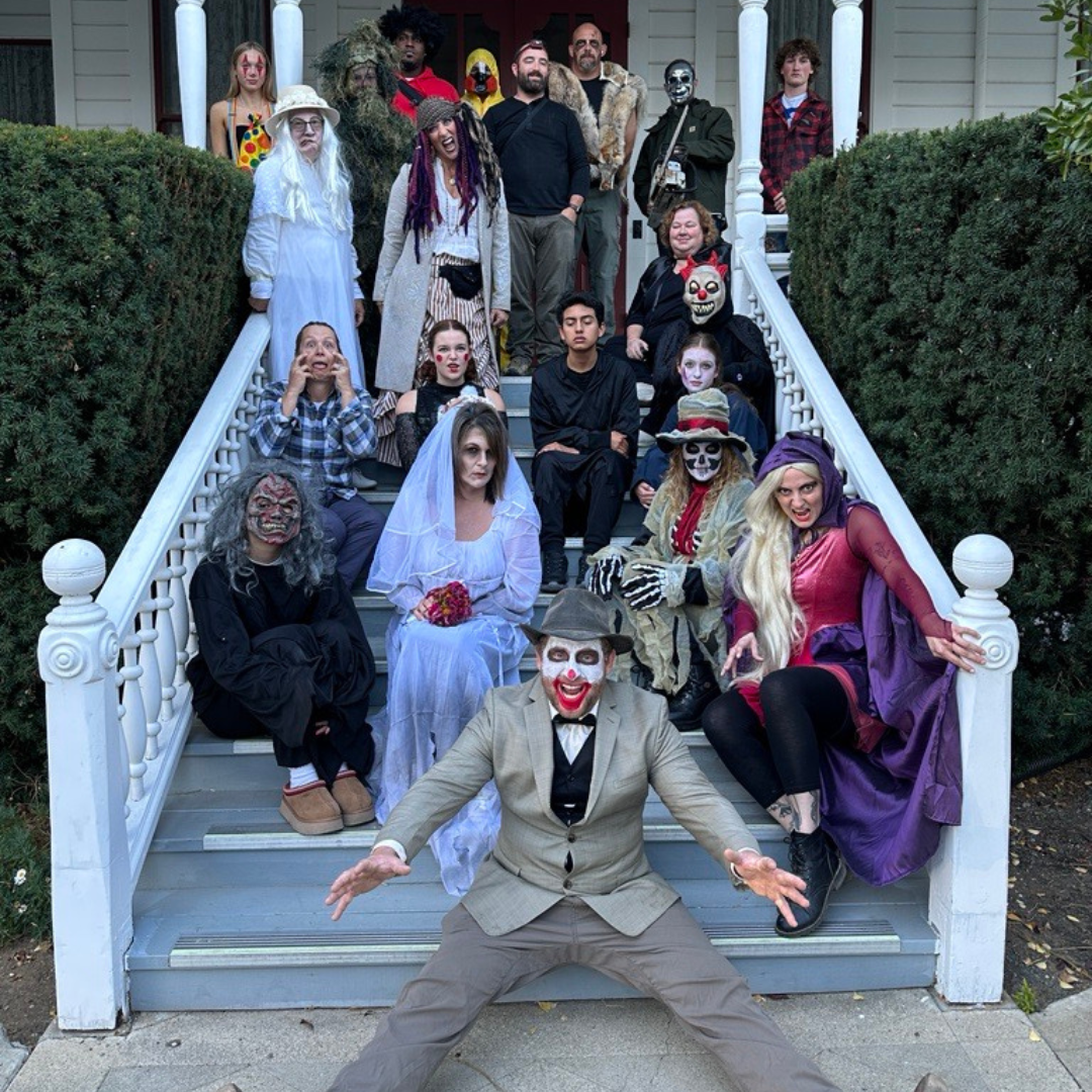 A large group in costumes, including ghosts, clowns, and zombies, poses on the stairs of a house for a Halloween-themed photo.