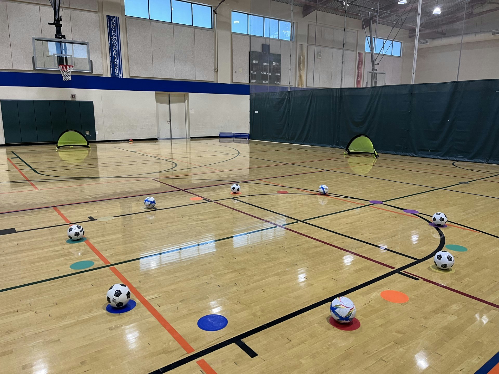 An indoor basketball court set up for a children's soccer activity with balls and colorful cones.
