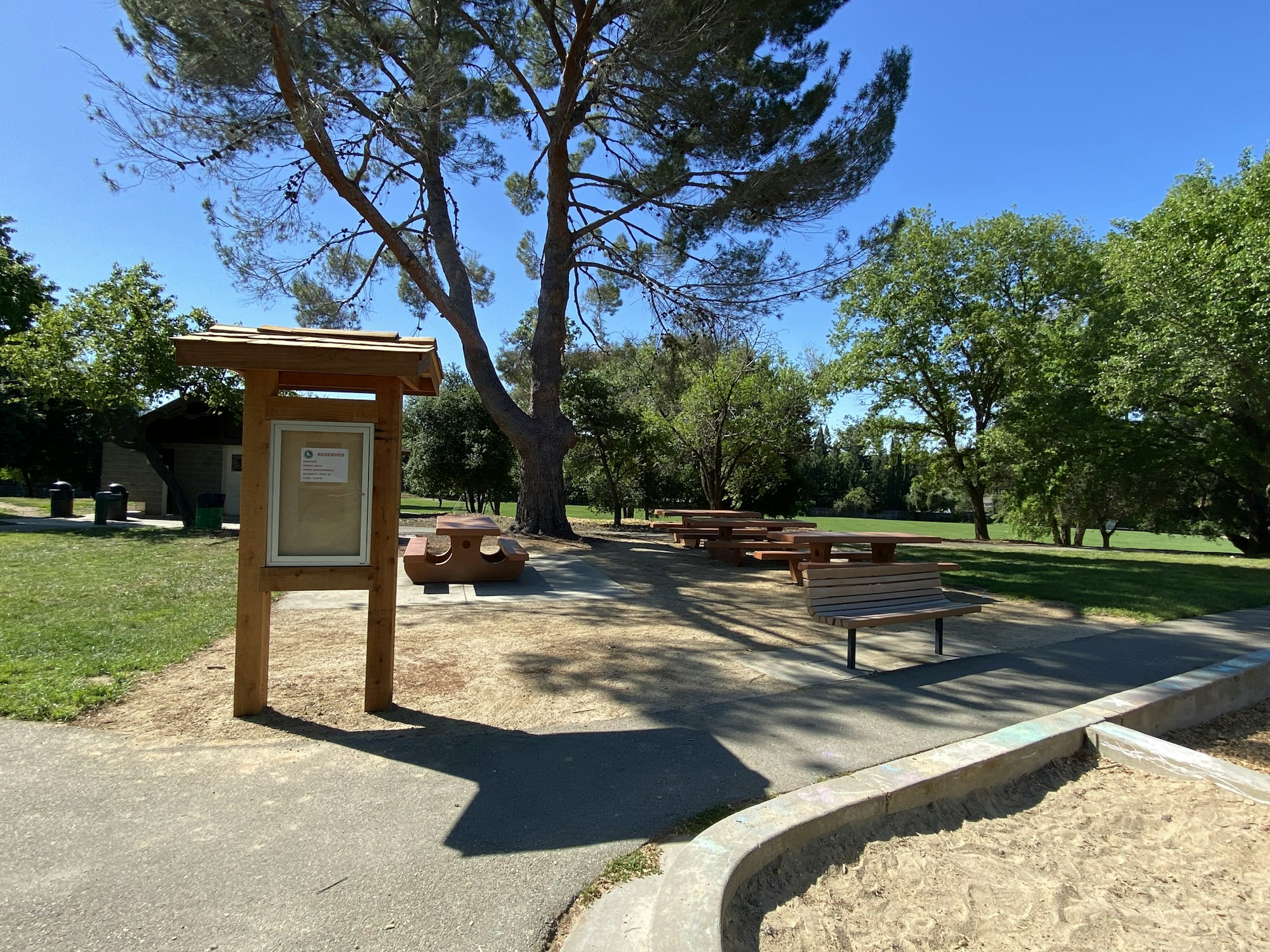 A sunny park scene with an information board, benches, trees, and a sandbox.