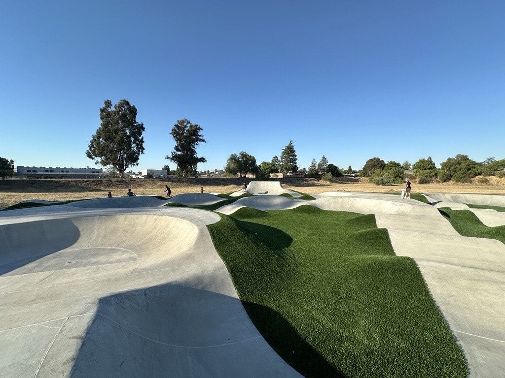 A modern skatepark features smooth concrete ramps and green turf elements under a clear blue sky, with people enjoying the space.