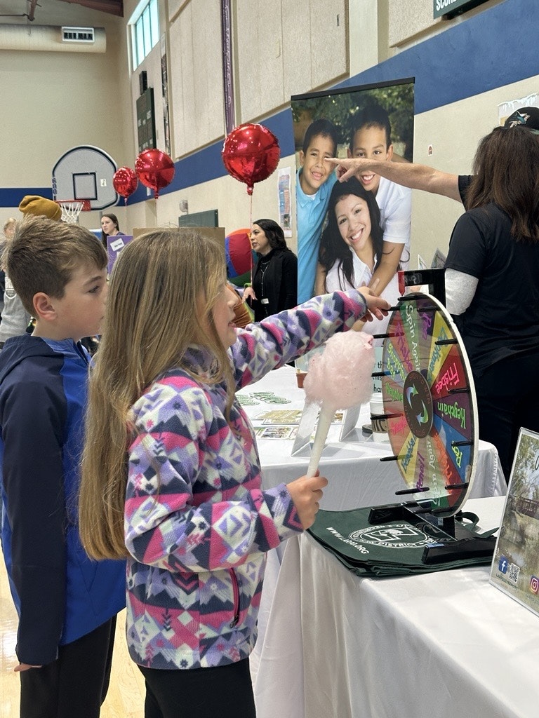 A girl spins a colorful wheel while holding cotton candy, with a boy next to her at a lively indoor event. Red balloons decorate the space.
