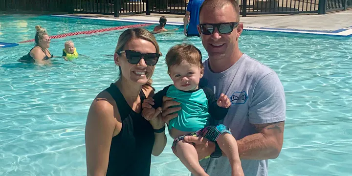 A family enjoys a sunny day at the pool, posing together with a toddler wearing a swim outfit. Happy vibes all around!