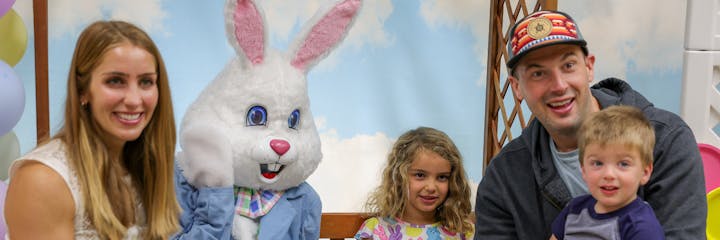 A family with children poses with a person in an Easter Bunny costume, smiling in a festive setting with balloons.