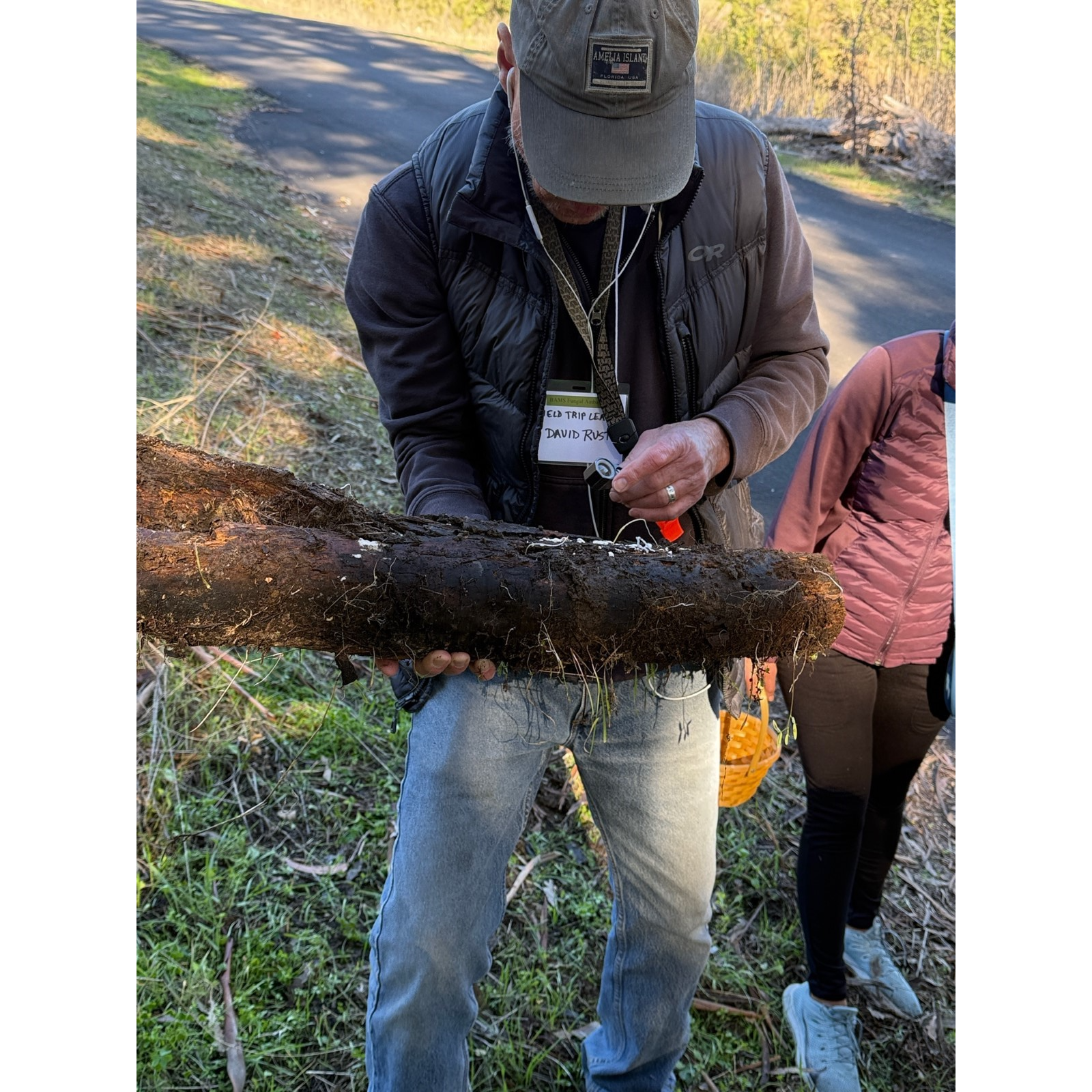 A person examines a log outdoors while wearing casual clothing and a cap.