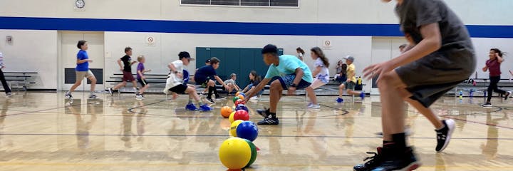 Kids playing dodgeball in a gym, running and picking up colorful balls from the floor.