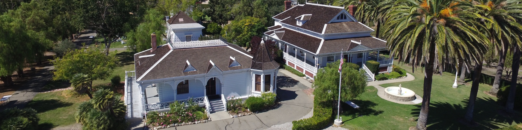 Two Victorian-style houses with brown roofs surrounded by palm trees and a garden with a flagpole and fountain.