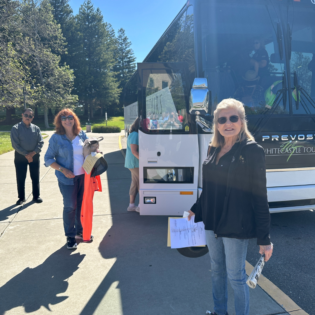 A group of people standing near a tour bus, some holding papers, with trees in the background.