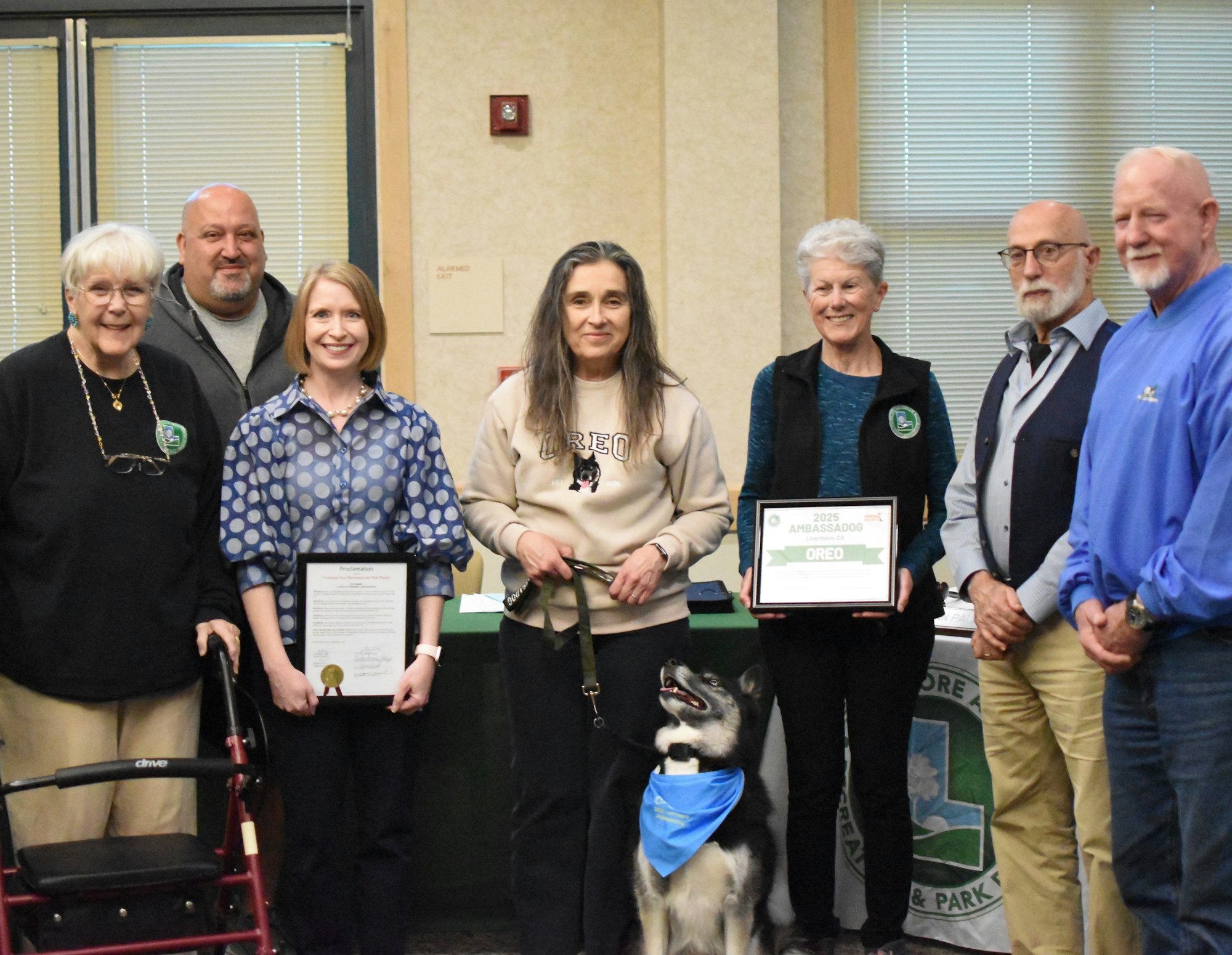 Seven people and a dog posing indoors, with two people holding certificates.